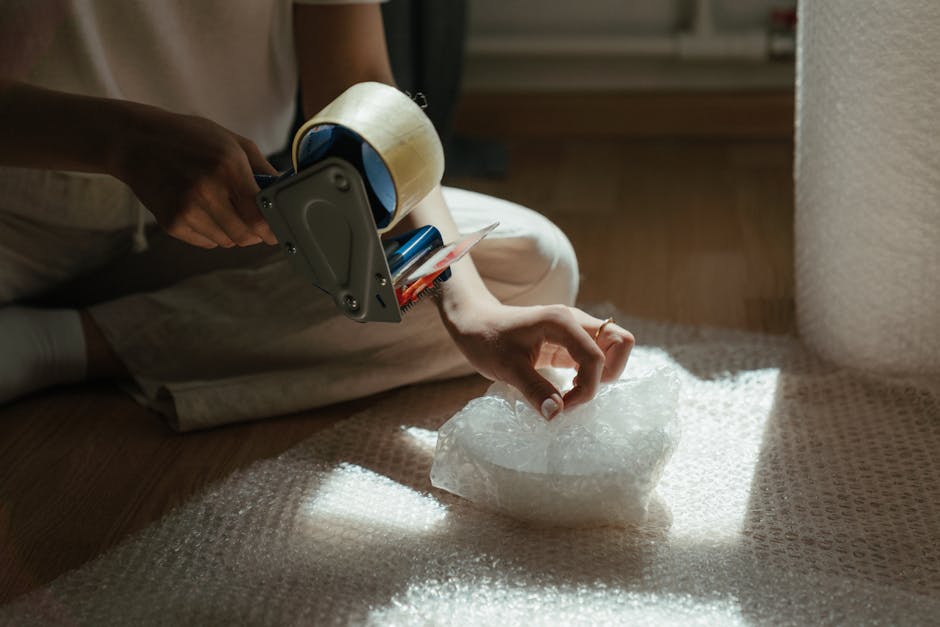 A person seated on the floor inside a home, using their left hand to carefully handle a piece of bubble wrap, which is placed on a textured beige carpet. The individual is wearing light-colored clothing, and their right hand holds a packing tape dispenser with clear packing tape, possibly preparing fragile items for transport. Nearby, a cardboard box is partially visible, indicating packing and moving preparations. The scene includes a beige upholstered piece of furniture on the right side and a wooden floor with soft natural lighting illuminating the area, reflecting typical home relocation activities. The image showcases the careful handling and packing process involved in home removals, emphasizing the importance of proper packaging for delicate items, as performed by [COMPANY_NAME], a professional removals service specializing in furniture transport and packing for house relocations.