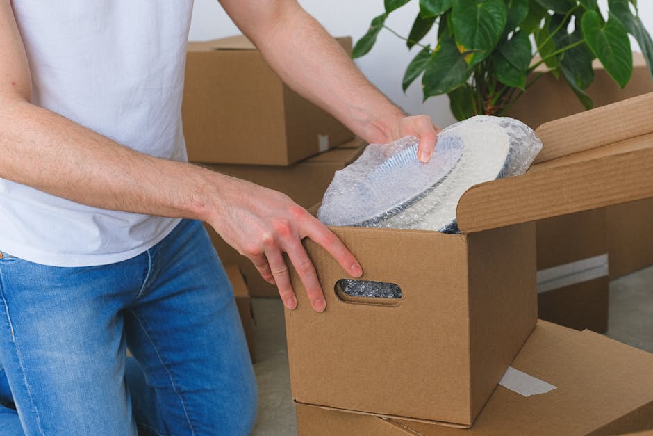 A person wearing a white t-shirt and blue jeans is seen packing a fragile item, possibly a ceramic or glass plate, into a cardboard box with cut-out handles. The item is wrapped in bubble wrap for protection. The setting appears to be inside a home, with other unpacked boxes and packing materials visible in the background, including a large green plant near the corner. The person is carefully placing the item into the box, which is positioned on a flat surface in preparation for a house move, with a focus on proper packing and handling for furniture transport and home relocation. This scene demonstrates the packing process in a professional removals context, emphasizing careful handling of delicate objects during the loading process, as part of a comprehensive moving service by Man With a Van Queensbury, supporting safe and efficient logistics for local moves.