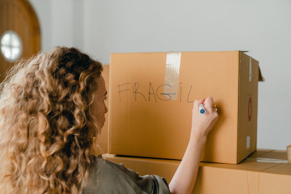 A person seated on the floor inside a home, using their left hand to carefully handle a piece of bubble wrap, which is placed on a textured beige carpet. The individual is wearing light-colored clothing, and their right hand holds a packing tape dispenser with clear packing tape, possibly preparing fragile items for transport. Nearby, a cardboard box is partially visible, indicating packing and moving preparations. The scene includes a beige upholstered piece of furniture on the right side and a wooden floor with soft natural lighting illuminating the area, reflecting typical home relocation activities. The image showcases the careful handling and packing process involved in home removals, emphasizing the importance of proper packaging for delicate items, as performed by [COMPANY_NAME], a professional removals service specializing in furniture transport and packing for house relocations.