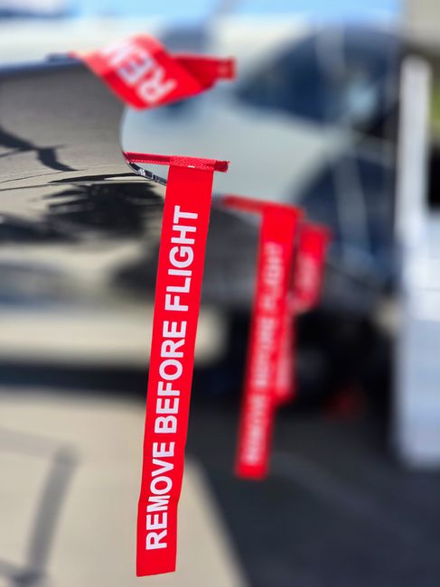 A close-up view of a dark grey van with red and white warning tape hanging from its side, displaying the message 'REMOVE BEFORE FLIGHT' in bold white letters. The tape is attached near the vehicle's door handle and drapes downward, partially obscuring the van's surface. The background includes a blurred urban setting with other vehicles and buildings, suggesting the van is parked outside a residential property. Inside the property, cardboard boxes, some wrapped in clear plastic, are placed near the entrance, indicating preparations for a house relocation or furniture transport. An open doorway is visible, with the interior lighting highlighting stacked boxes and household items ready to be moved, in alignment with professional packing and moving services offered by Man With a Van Queensbury as part of their removals process.