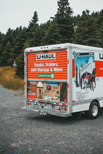 The image shows the rear view of a white U-Haul moving truck parked on a gravel surface beside a rural road. The truck's back doors are partially open, revealing a glimpse of packed cardboard boxes, some with printed labels, and other packing materials such as bubble wrap and protective foam inside. The truck features bright orange and blue branding, advertising trucks, trailers, self-storage, and moving services, with the U-Haul logo and website visible. Behind the truck, there is a backdrop of dense trees and greenery under an overcast sky, suggesting a countryside setting. The scene is representative of home relocation or furniture transport activities, aligning with professional removals and moving services like those provided by Man With a Van Queensbury, focusing on packing, loading, and transportation aspects of house moves.