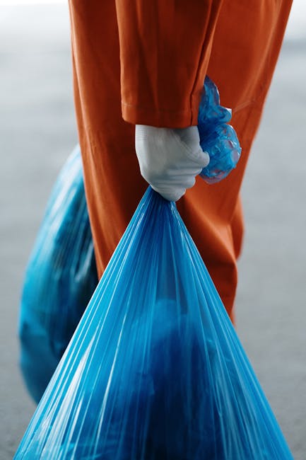 In the image, a person dressed in an orange jumpsuit is holding a blue plastic rubbish bag in their right hand, which is covered with a white glove. The individual is standing outdoors on a paved surface, possibly near a property or loading area. Nearby, there are additional blue bags, suggesting a collection of waste or bulky items for removal as part of a house clearance or home relocation process. The environment appears well-lit, with natural daylight illuminating the scene. This image visually supports services related to house removals, furniture transport, or rubbish clearance, which Man With a Van Queensbury might provide to assist with packing, waste collection, or moving logistics while maintaining a professional, neutral tone appropriate for the topic “Bulky waste in Queensbury: Collection or removal?”