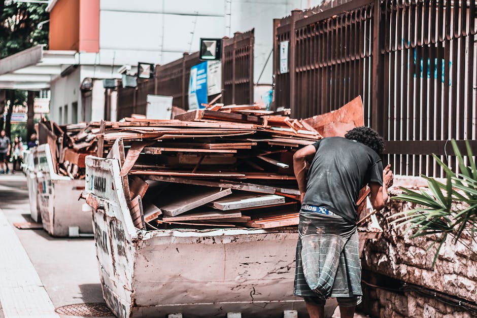 A person wearing a dark T-shirt and plaid shorts is standing outside a property near a large, open skip filled with construction debris, including wooden planks, tiles, and plastic wrapping materials. The skip is positioned on a paved sidewalk adjacent to a high brown metal fence and a building with a white exterior. In the background, there are signs, a few pedestrians, and trees. The scene appears to be part of a house clearance or renovation project, involving the loading or removal of bulky waste items, which is relevant to house removals services offered by Man With a Van Queensbury, particularly during packing and moving logistics or bulky waste collection.