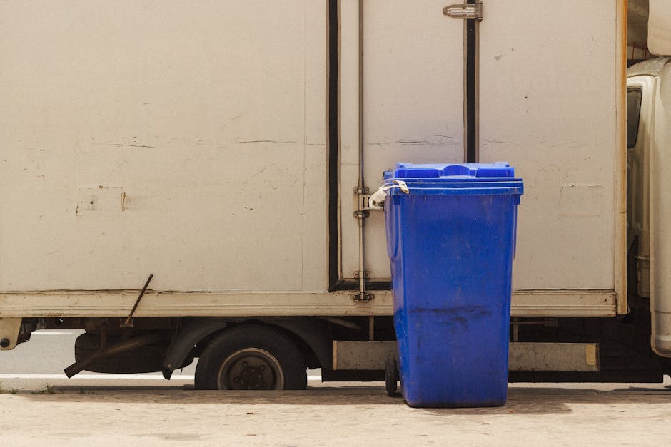 In the image, a person dressed in an orange jumpsuit is holding a blue plastic rubbish bag in their right hand, which is covered with a white glove. The individual is standing outdoors on a paved surface, possibly near a property or loading area. Nearby, there are additional blue bags, suggesting a collection of waste or bulky items for removal as part of a house clearance or home relocation process. The environment appears well-lit, with natural daylight illuminating the scene. This image visually supports services related to house removals, furniture transport, or rubbish clearance, which Man With a Van Queensbury might provide to assist with packing, waste collection, or moving logistics while maintaining a professional, neutral tone appropriate for the topic “Bulky waste in Queensbury: Collection or removal?”