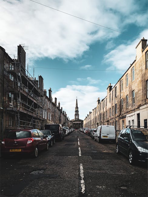 An image of a narrow residential street with parked cars lining both sides on a cloudy day. The street is paved and slightly uneven, with trees on the left side displaying autumn foliage in shades of 