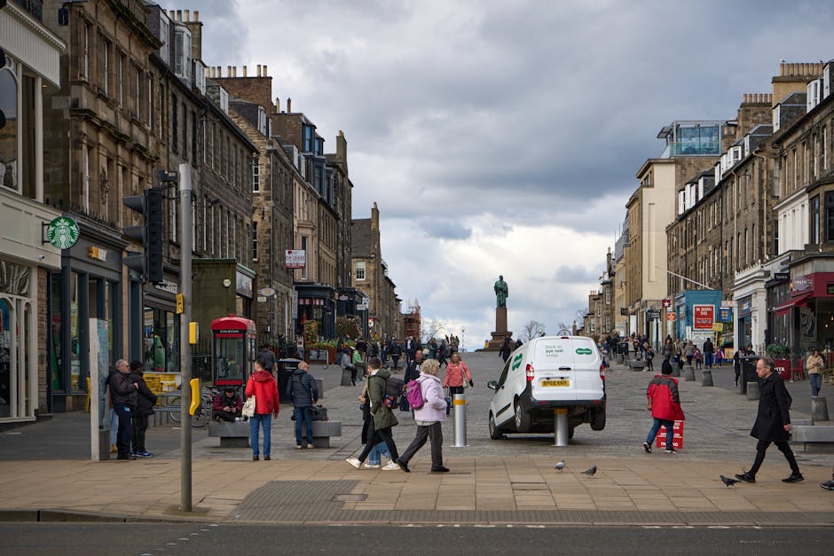 A busy street scene in Queensbury with pedestrians walking along the pavement, some carrying bags or backpacks, and a white delivery van parked near the curb supported by small bollards. In the background, multiple multi-storey historic buildings line the street, featuring traditional brickwork and large windows, with a statue of a seated figure atop a pedestal standing at the far end of the street. The sidewalk is partially set back with a few street furniture elements, including a red telephone booth and a green Starbucks coffee sign attached to one of the shopfronts. Overhead, grey clouds fill the sky, indicating overcast weather. This scene depicts the typical environment for home relocation and furniture transport services in Queensbury, with the van positioned for loading or unloading furniture and moving boxes, and pedestrians observed in the midst of their daily activities, supporting the context of a town centre suitable for house removals facilitated by companies like Man With a Van Queensbury.