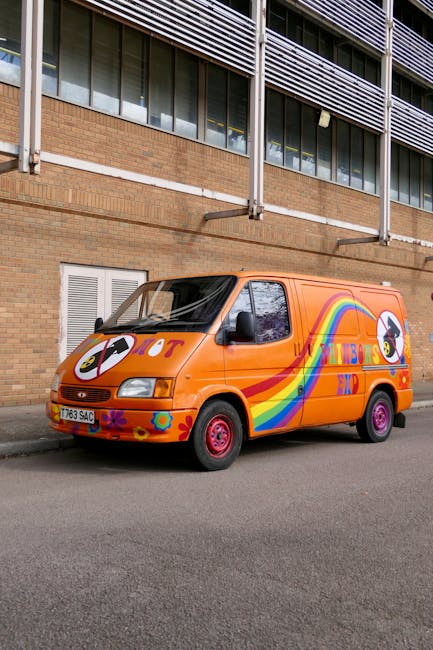 An orange van with rainbow and unicorn graphics parked on a city street in front of a brick building with multiple windows and external metal blinds. The van's front windshield and side windows are visible, and it has red wheels. The pavement is along the curb, and the scene suggests a moving or transportation context, consistent with house removals or furniture transport services offered by Man With a Van Queensbury. The environment is well-lit, and the van is positioned for loading or unloading, possibly as part of a home relocation process involving packing boxes, furniture, or appliances nearby.