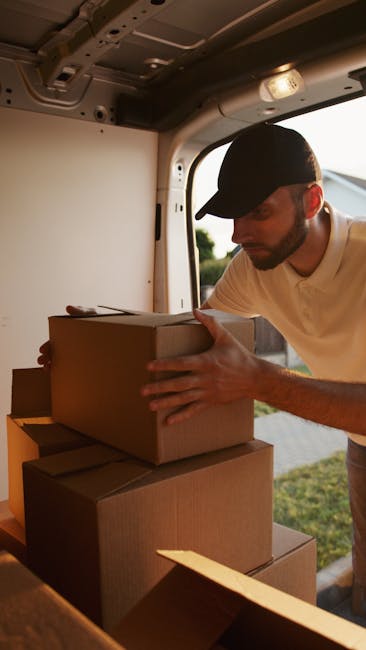 A man wearing a black cap and a light-colored polo shirt is inside a guided van, carefully handling and stacking three cardboard boxes on top of each other as part of a home relocation process. The interior of the van is illuminated by warm light, with visible ceiling fixtures and part of the side window showing an outdoor scene with greenery and sunlight. The boxes are plain, sealed with packing tape, and are being arranged for efficient loading, reflecting a professional furniture transport and packing process associated with local house removals in the Queensbury area. This is part of a typical loading process managed by Man With a Van Queensbury, a company specialising in removals and moving logistics.