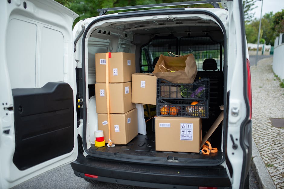 The rear of a white van, belonging to Man With a Van Queensbury, is open and packed with various moving supplies including multiple cardboard boxes of different sizes, some sealed with tape and labeled with barcodes, a small paper bag, a black plastic crate filled with oranges and other produce, and a cardboard box marked with fragile symbols. The boxes are arranged to maximize space inside the van, which is situated outdoors on a paved area near a residential street with a cobblestone pavement and a white fence visible in the background. A black and orange ratchet strap, along with other packing materials, are also visible inside the vehicle. The scene captures the loading process involved in home relocation or furniture transport, with natural daylight illuminating the space, highlighting the packing and logistical steps typical of a professional removals service such as [COMPANY_NAME], in preparation for a local move from Kenton to Queensbury.