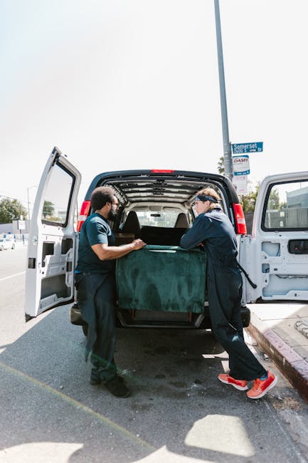 A man wearing a black cap and a light-colored polo shirt is inside a guided van, carefully handling and stacking three cardboard boxes on top of each other as part of a home relocation process. The interior of the van is illuminated by warm light, with visible ceiling fixtures and part of the side window showing an outdoor scene with greenery and sunlight. The boxes are plain, sealed with packing tape, and are being arranged for efficient loading, reflecting a professional furniture transport and packing process associated with local house removals in the Queensbury area. This is part of a typical loading process managed by Man With a Van Queensbury, a company specialising in removals and moving logistics.