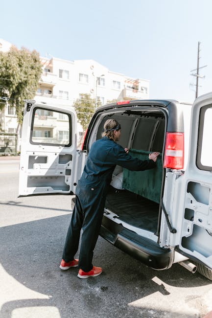 A man with a beard and short hair, dressed in a white shirt, black t-shirt, grey shorts, and white sneakers with black stripes, sitting on the side of a white moving van with its side door open. He is