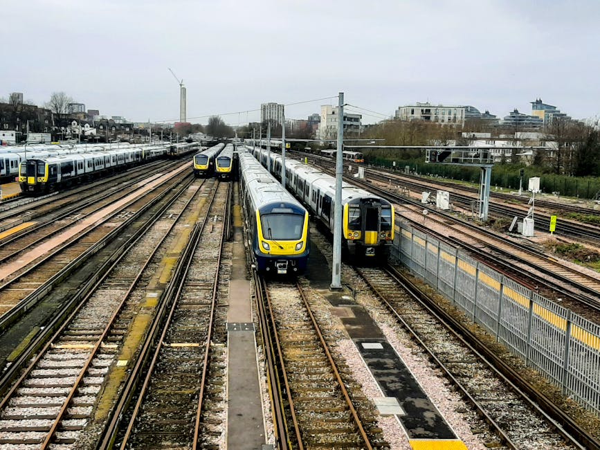 A multi-track railway station with several modern passenger trains parked on the tracks, featuring yellow and grey front panels. The trains are positioned parallel to each other, with some trains on the left side of the image and others closer to the centre. Overhead electric wires and supporting poles run above the tracks. In the background, there are various urban buildings, including high-rise and mid-rise structures, with some trees and greenery visible beyond the station. The scene appears to be overcast, with grey clouds covering the sky. The station's platform is not directly visible, but safety barriers and fencing along the tracks are evident. The environment suggests a busy transportation hub, potentially during a home relocation or commuting period. Although primarily focused on rail transport, the image's context aligns with logistics and moving services provided by a company like Man With a Van Queensbury, highlighting infrastructure relevant to moving and professional transportation.