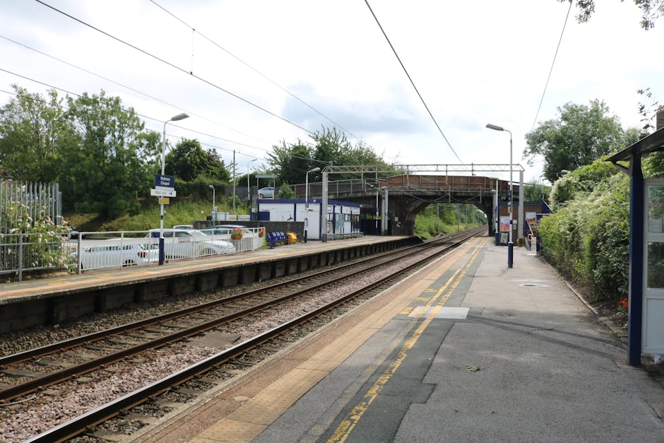 An empty train station platform in Queensbury with parallel railway tracks running through the centre, overhead electric wires and signals visible above the tracks, a small white building with blue signage near the platform edge, and a covered shelter on the right side of the platform. The station is surrounded by lush green bushes and trees, with street lamps mounted on poles along the platform. The pathway on the platform features tactile paving strips for accessibility, and a fence separates the station from a parking lot with several parked cars. Overhead, there is a bridge for pedestrians and vehicles, constructed of metal and brick, providing an overpass for crossing the railway. The sky is partly cloudy with patches of sunshine illuminating the scene, suggesting a typical day for home relocation or furniture transport involving careful loading and moving logistics facilitated by [COMPANY_NAME].