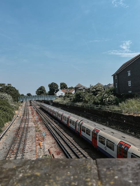 A multi-track railway station with several modern passenger trains parked on the tracks, featuring yellow and grey front panels. The trains are positioned parallel to each other, with some trains on the left side of the image and others closer to the centre. Overhead electric wires and supporting poles run above the tracks. In the background, there are various urban buildings, including high-rise and mid-rise structures, with some trees and greenery visible beyond the station. The scene appears to be overcast, with grey clouds covering the sky. The station's platform is not directly visible, but safety barriers and fencing along the tracks are evident. The environment suggests a busy transportation hub, potentially during a home relocation or commuting period. Although primarily focused on rail transport, the image's context aligns with logistics and moving services provided by a company like Man With a Van Queensbury, highlighting infrastructure relevant to moving and professional transportation.