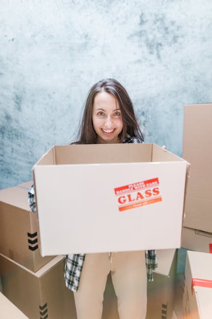 A young woman with long brown hair smiling as she holds an empty cardboard box labeled 'fragile glass' inside a room with a plain, textured light blue wall. She is dressed in a casual plaid shirt over light-colored pants. Around her are several other cardboard boxes, some sealed with packing tape and others open, indicating an ongoing home relocation or packing process. The boxes are made of brown corrugated cardboard, and some have black and red tape or printed markings. The scene is lit with natural or soft artificial lighting, capturing a moment of packing preparation for a house move. The image reflects the typical activities involved in furniture transport and packing during moving services, highlighting the logistical aspect of home relocation handled by companies like Man With a Van Queensbury.