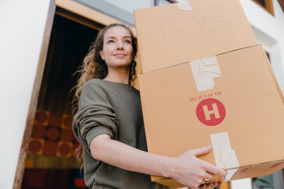 A woman with curly brown hair, wearing a grey long-sleeved sweatshirt, is holding two large cardboard moving boxes inside a home, near an open doorway with a wooden frame. One box has 'toys' written on it in pencil, and both boxes are secured with packing tape and reinforced with white straps. These boxes are part of a home relocation process, likely prepared for transport or loading onto a van. The background includes interior decor with visible fabric and wood elements, and natural lighting illuminates the scene, emphasizing the careful handling of packing materials involved in furniture transport and packing and moving services. Man With a Van Queensbury specializes in home removals, including packing and loading, supporting customers in efficient house moves.