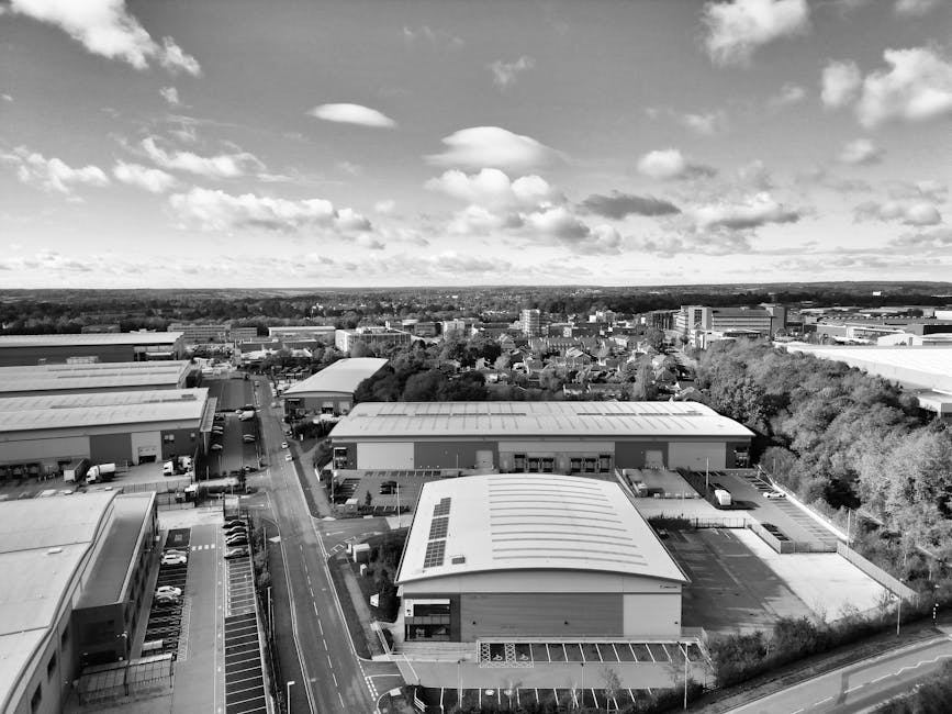 An aerial view in black and white showing a cityscape with multiple industrial warehouses featuring flat, slightly curved roofs, some of which have solar panels installed. The warehouses are situated along a main road with parked vehicles and trucks on the premises, indicating logistics activity related to house removals and furniture transport. To the right, there are trees forming a green boundary, while in the background, a mix of residential and commercial buildings extends towards the horizon under partly cloudy skies. The scene captures the loading area of a warehouse or storage facility, likely used by Man With a Van Queensbury for packing, loading, and coordinating home relocation and moving services. The perspective emphasizes the physical logistics involved in furniture transport and professional removals, with the environment well-lit and in focus, representing an active site of moving-related operations.