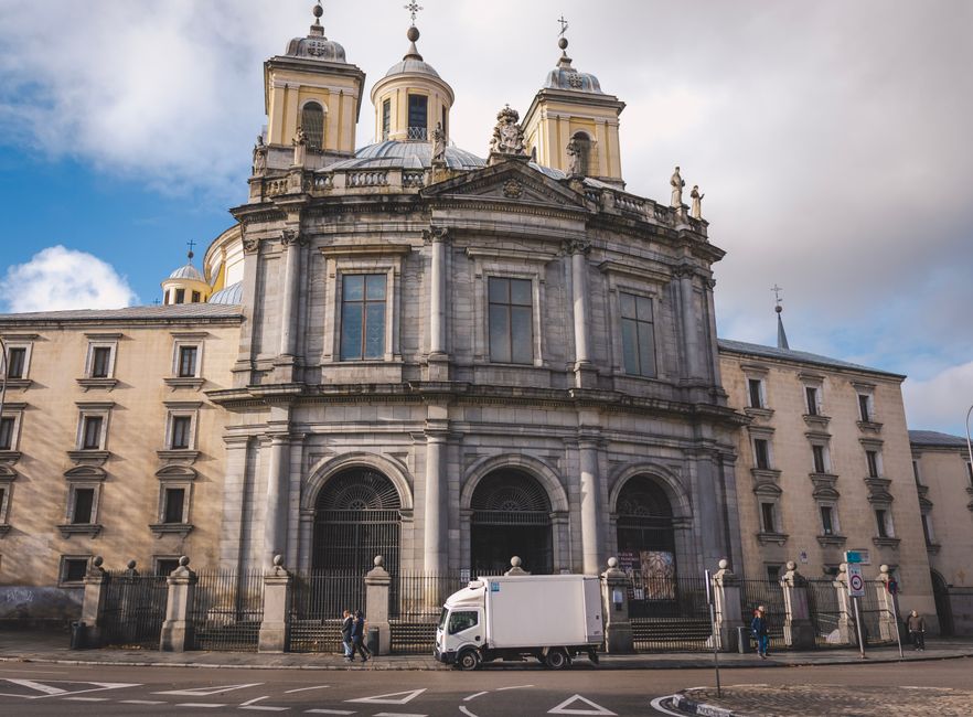 A white moving van from Man With a Van Queensbury is parked on the street in front of a historic baroque-style building with large arched windows, ornate stonework, and decorative statues on the roof. The van is positioned close to the building's entrance, which is surrounded by a black wrought iron fence. Street pedestrians are walking nearby, some passing by the vehicle on the sidewalk, while the driver appears to be engaged in loading or unloading furniture and boxes. The interior of the van is not visible, but it is part of the home relocation or furniture transport process managed by Man With a Van Queensbury, featuring a large cargo area suitable for packing and moving household items. The scene is illuminated by natural daylight, with partly cloudy skies overhead, reflecting typical urban moving logistics and the loading process during a house or office move.