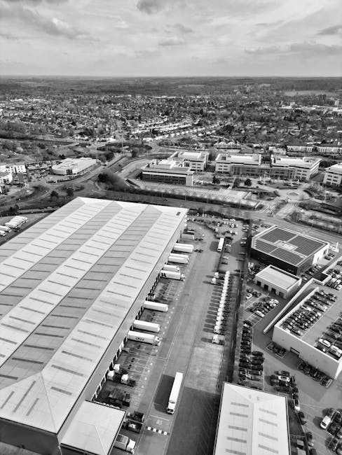 An aerial view in black and white showing a cityscape with multiple industrial warehouses featuring flat, slightly curved roofs, some of which have solar panels installed. The warehouses are situated along a main road with parked vehicles and trucks on the premises, indicating logistics activity related to house removals and furniture transport. To the right, there are trees forming a green boundary, while in the background, a mix of residential and commercial buildings extends towards the horizon under partly cloudy skies. The scene captures the loading area of a warehouse or storage facility, likely used by Man With a Van Queensbury for packing, loading, and coordinating home relocation and moving services. The perspective emphasizes the physical logistics involved in furniture transport and professional removals, with the environment well-lit and in focus, representing an active site of moving-related operations.