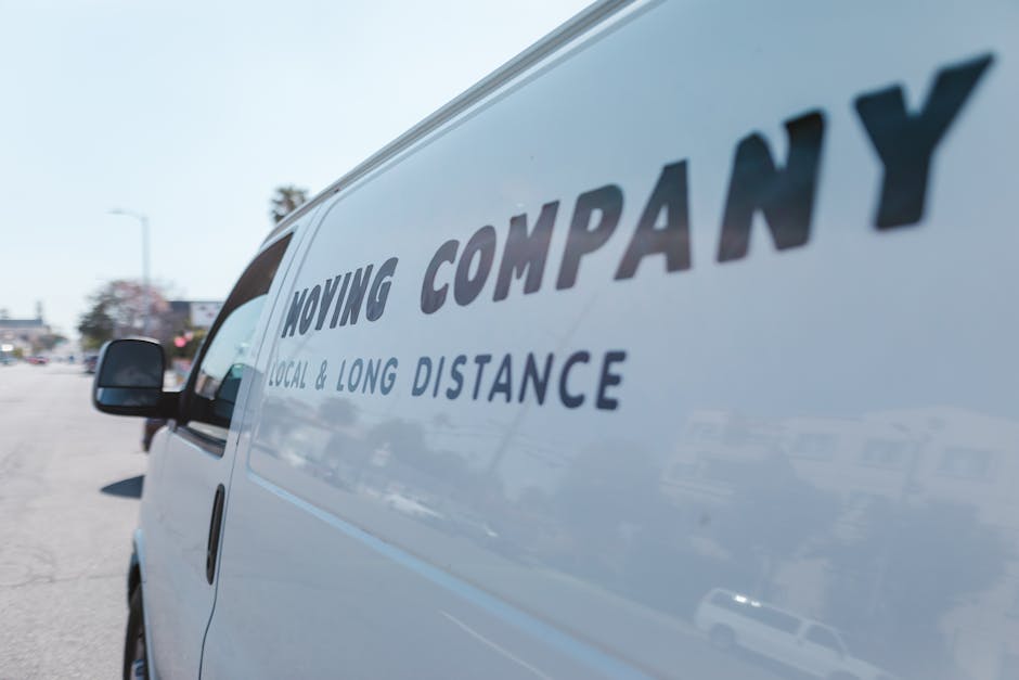 Close-up view of a white moving van parked outdoors in a parking lot, with the side panel displaying the company logo and text 'Moving Company' and 'Local & Long Distance.' The van's side window is partially visible, and reflections of nearby cars and buildings can be seen on the vehicle's surface. In the background, blurred images of other parked vehicles, street lamps, and trees are visible under bright daylight, indicating an environment suitable for home relocation or furniture transport services. The scene captures the loading area near the van, suggesting preparation for a house move or packing and moving operations, with the focus on professional vehicle engagement in the relocation process, as associated with [COMPANY_NAME]'s removals services, including last-minute and urgent moves in Queensbury.