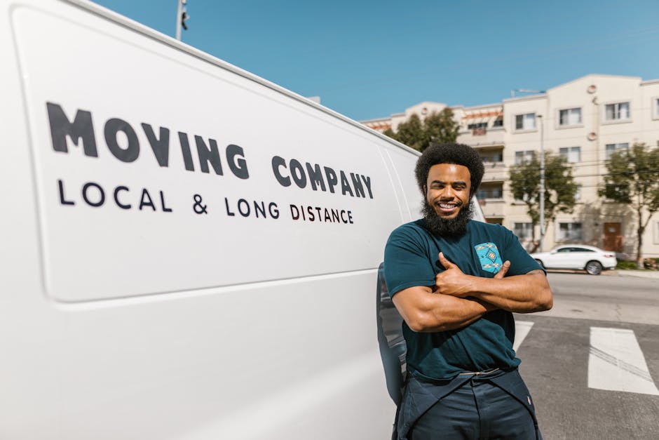 Close-up view of a white moving van parked outdoors in a parking lot, with the side panel displaying the company logo and text 'Moving Company' and 'Local & Long Distance.' The van's side window is partially visible, and reflections of nearby cars and buildings can be seen on the vehicle's surface. In the background, blurred images of other parked vehicles, street lamps, and trees are visible under bright daylight, indicating an environment suitable for home relocation or furniture transport services. The scene captures the loading area near the van, suggesting preparation for a house move or packing and moving operations, with the focus on professional vehicle engagement in the relocation process, as associated with [COMPANY_NAME]'s removals services, including last-minute and urgent moves in Queensbury.